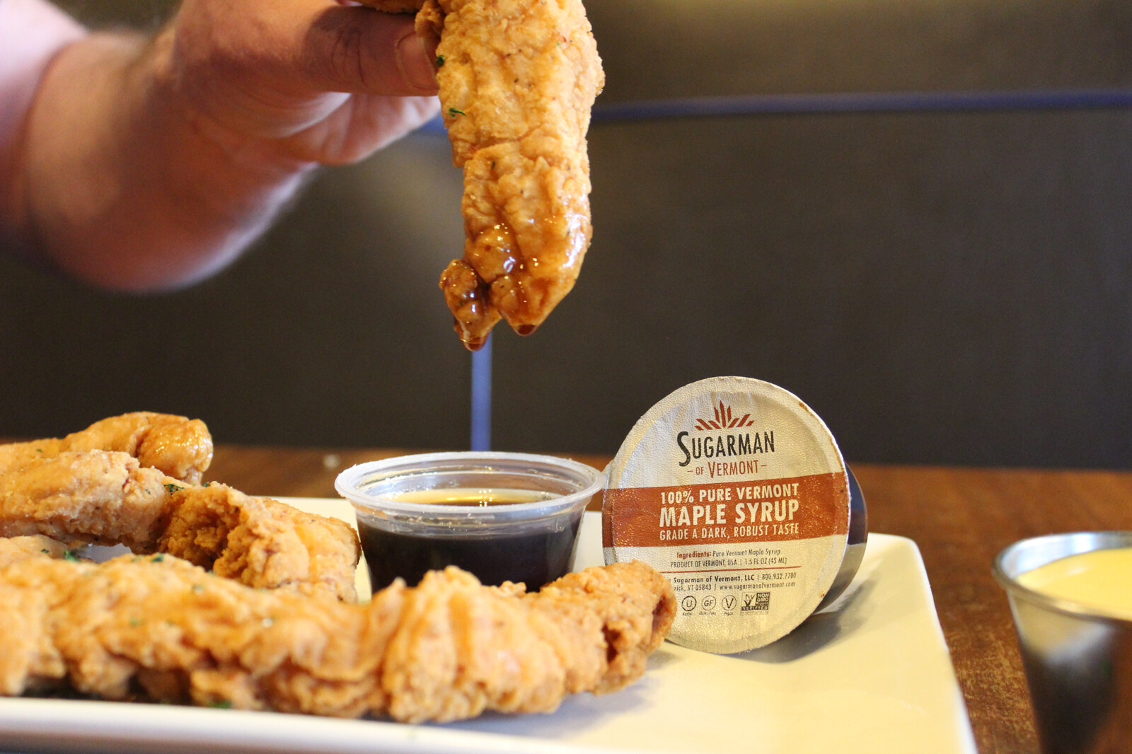 A hand dips fried chicken into a cup of maple syrup on a plate.