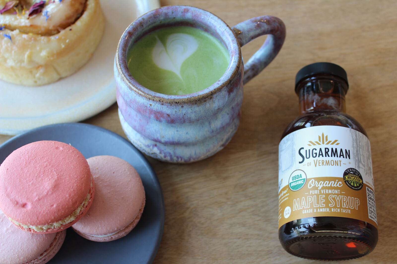 A colorful mug with matcha, macaroons, a pastry, and a bottle of maple syrup.