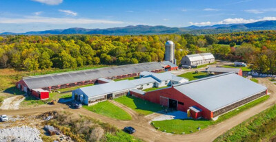 Aerial view of a farm with red barns, silos, and surrounding colorful trees and mountains.