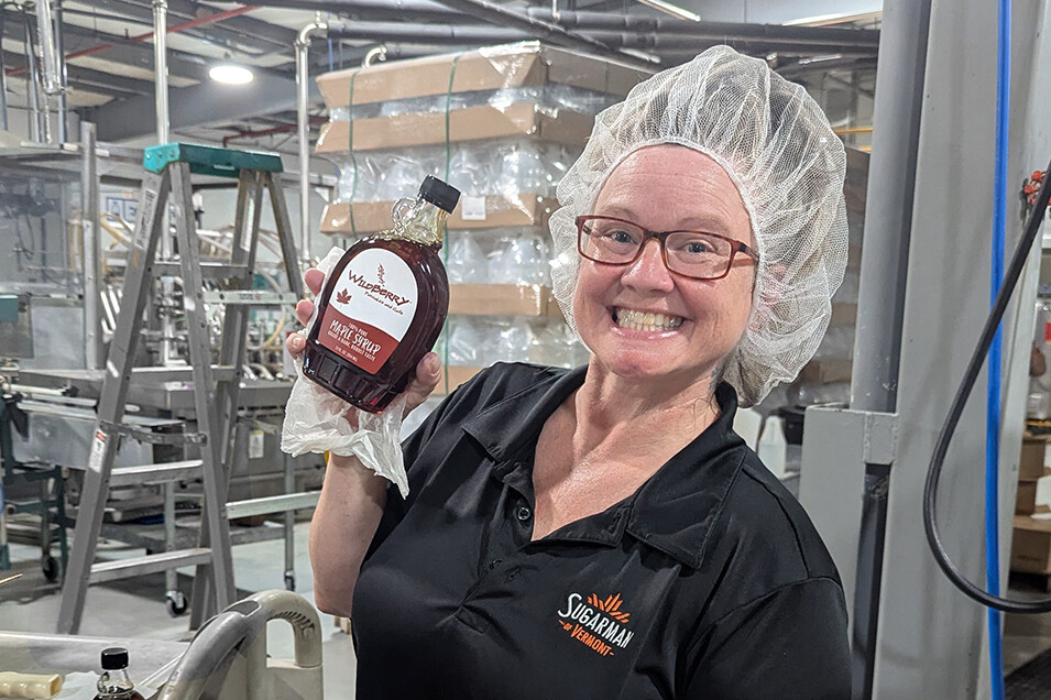 Smiling woman in a hairnet holds a bottle of maple syrup in a production facility.