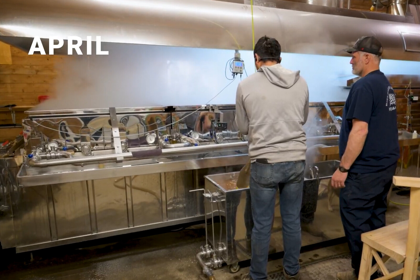 Two people work with a maple syrup production setup, steam rising around them.