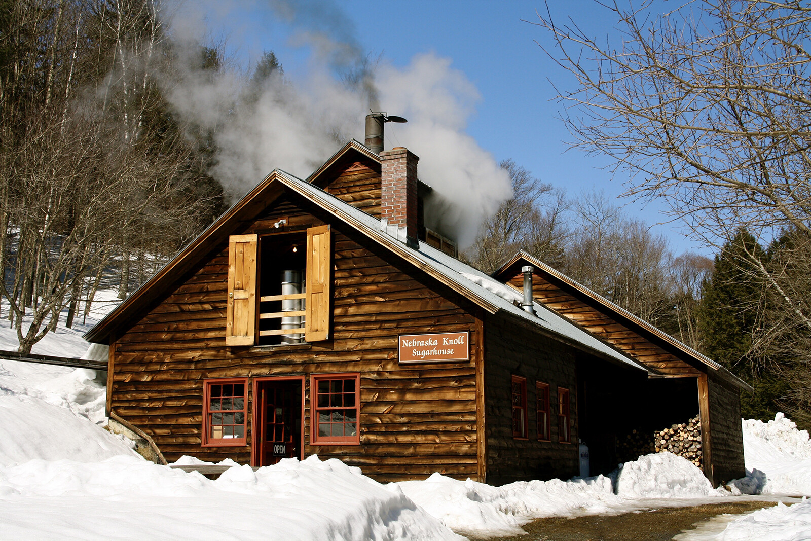 A wooden sugarhouse with smoke rising, surrounded by snow and trees.