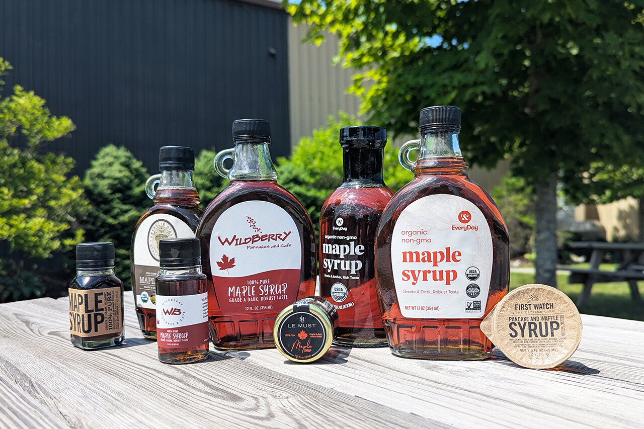 Assorted bottles of maple syrup arranged on a wooden table with greenery in the background.