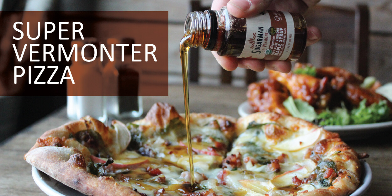 A person drizzles syrup on a Super Vermonter pizza, with chicken wings in the background.