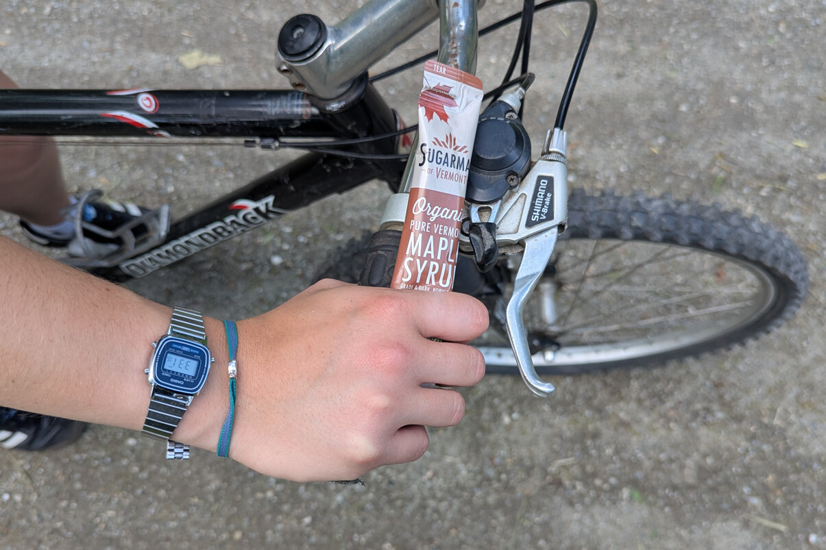 A hand holding a tube of maple syrup while resting on a bicycle handlebar.